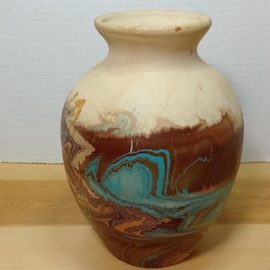 Puede incluir: Un jarrón decorativo de cerámica con la parte superior de color crema y un cuerpo con diseños marrones y turquesas. El jarrón tiene forma redondeada con cuello estrecho y base ancha. Los colores se mezclan en un efecto veteado.