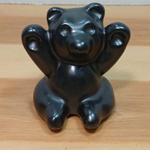 May include: A black bear figurine with its arms raised. The bear is glossy and appears to be made of a solid material. The figurine is sitting on a wooden surface. The image was taken on Nov 18, 2025, at 9:54 AM.