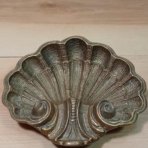 May include: A brass dish shaped like a scallop shell with a textured, ribbed surface and two prominent curled decorative elements at the base. The metal has a dark, aged patina with highlights along the raised ridges of the shell design.