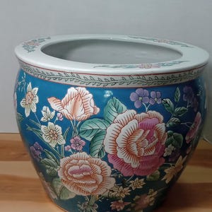Könnte beinhalten: Großer, runder Keramik-Blumentopf mit breitem Rand. Der Topf ist teal mit einem Blumendesign mit rosa, pfirsichfarbenen und weißen Rosen, zusammen mit anderen Blumen und grünen Blättern. Der Rand ist weiß mit einem dekorativen Rand.