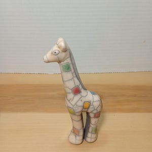 Puede incluir: Una figura de jirafa de cerámica de color crema con un cuello largo. La jirafa tiene parches cuadrados de colores rojo, amarillo, verde y azul. La figura está sobre una superficie de madera. El texto "shot on moto g stylus (2023)" y "Themadhaggler" son visibles.