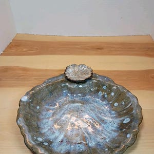 Puede incluir: Un plato de cerámica redondo con borde festoneado y un esmalte brillante y moteado en tonos marrón, azul y blanco. Un pequeño adorno texturizado en forma de flor está unido al borde superior del plato, con un acabado en beige claro y marrón.