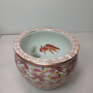May include: A round porcelain bowl featuring a hand-painted orange goldfish and green aquatic plants inside. The exterior is decorated with a pink and white lattice pattern, floral motifs, and gold trim along the rim and the lattice intersections.