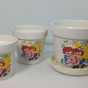 Peut inclure: Trois pots de fleurs en céramique blanc cassé de tailles différentes, chacun orné d'un motif coloré représentant deux poupées aux cheveux roux assises sous un arbre jaune. Le plus grand pot possède une soucoupe circulaire assortie.