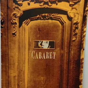 Puede incluir: Un cartel de la película "Cabaret" vintage. El cartel presenta una ilustración detallada de una puerta de madera con tallas ornamentadas. La palabra "CABARET" se muestra en letras doradas, con una pequeña imagen de un rostro encima.