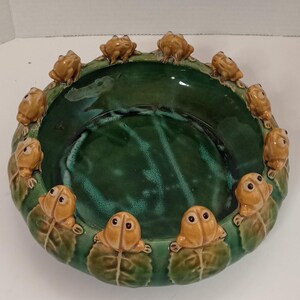 May include: A ceramic dish featuring a deep green interior and a rim adorned with small, tan frog figurines. The dish is encircled by green lily pads, creating a whimsical, nature-inspired design. The dish is approximately 25 cm in diameter.