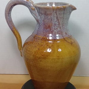 May include: A handcrafted ceramic pitcher with a curved handle and a spout. The pitcher features a gradient of colors, transitioning from a golden brown base to a purple and gray rim. It sits on a black circular base.