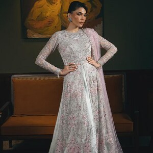 Pode incluir: Um vestido longo cinza claro com intrincados bordados florais em rosa. O vestido apresenta mangas compridas e uma saia fluida, combinado com um xale rosa a condizer. A modelo está em frente a uma pintura e a um sofá castanho.