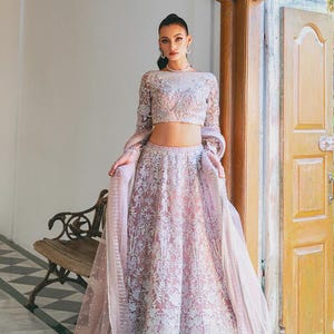 May include: A light pink Lehenga Choli, a traditional Indian outfit, featuring intricate embroidery. The ensemble includes a long-sleeved crop top and a flowing skirt, with a matching sheer dupatta. The text "STYLE - A" and "LEHENGA CHOLI" are visible.