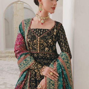 May include: A black and gold embroidered dress with long sleeves and a square neckline. The dress is adorned with gold embellishments and paired with a green and red shawl. The model is wearing gold jewellery, including a necklace, earrings, and rings. The text "STYLE 02" is in the top left corner.