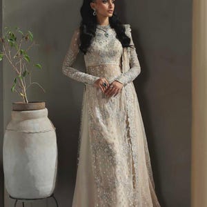 May include: An ivory-colored formal gown with intricate silver embroidery. The dress features long sleeves, a high neckline, and a flowing skirt with a sheer overlay. The model is wearing matching jewelry, including a headpiece and earrings.