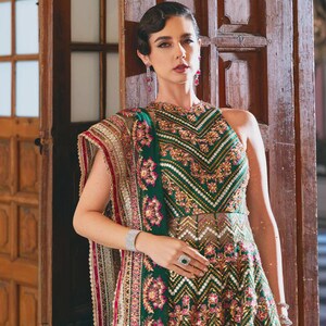 May include: A green and gold embroidered formal dress with a high neckline and a patterned design. The dress features a matching shawl with red and gold accents. The model is wearing a silver bracelet and ring.