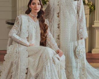 May include: Two women wearing matching white embroidered dresses. The dresses feature intricate lace detailing, long sleeves, and flowing skirts. One woman is seated, while the other stands, both posing in a well-lit room. The dresses are adorned with delicate embellishments.