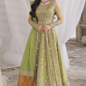 May include: An olive green formal gown with detailed embroidery and embellishments. The dress has long sleeves, a fitted bodice, and a flowing skirt. The gown features a detailed pattern with a contrasting orange and pink hemline. A matching scarf and necklace complete the look.