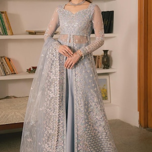 May include: A light blue formal gown with intricate embroidery and a sheer overlay. The gown features long sleeves, a high neckline, and a flowing skirt. The model is wearing a matching necklace and bracelet. The background includes bookshelves and a lamp.