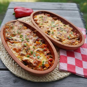 Może przedstawiać: Dwa owalne, terakotowe naczynia wypełnione zapiekanym daniem, posypane roztopionym serem i ziołami. Czerwona papryka i czerwono-biała serwetka w kratkę leżą na drewnianym stole.