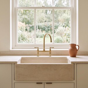 May include: A beige stone farmhouse sink with a brass bridge faucet featuring two handles. A terracotta pitcher sits on the light-colored countertop next to the sink. The sink is set into a cabinet unit with flat-front doors and metal pulls.