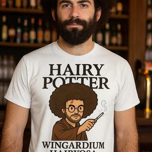 Puede incluir: Camiseta blanca con el texto "HAIRY POTTER" sobre una ilustración de dibujos animados de un hombre con afro, gafas y una varita. Debajo de la ilustración está el texto "WINGARDIUM HAIRYOSA" y el nombre de la marca.