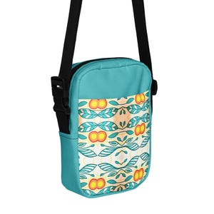 May include: A teal crossbody bag with a black adjustable strap. The bag features a white panel with a repeating pattern of orange, yellow, and teal leaf and flower designs. The bag has a small side pocket.