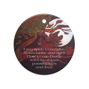 May include: A circular ornament featuring a dark background with a white moon and tree branches. The text reads: I am open to receive. Abundance and light flow to me freely with limitless possibilities and love. There is a small hole at the top.