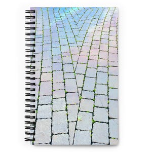May include: Spiral-bound notebook with a cover depicting a cobblestone pattern. The design features a light grey, textured surface with a network of rectangular stones, with green vegetation. The cover has a subtle rainbow effect.