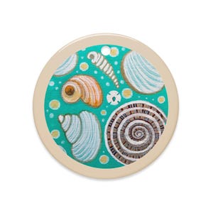 May include: Round ceramic ornament featuring a teal background with painted seashells in shades of orange, white, and brown. The ornament has a beige border and a hole at the top for hanging. The design evokes a coastal or beach theme.