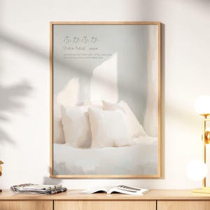 Puede incluir: Un estampado enmarcado con una imagen abstracta suave y clara de almohadas y una cama. El estampado tiene texto japonés en la parte superior. El estampado se exhibe sobre una mesa de madera con una lámpara y una escultura doradas.
