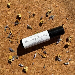 May include: A black and white essential oil roller bottle labeled "Dreamy Nights Restful Blend" lies on a textured brown surface, surrounded by dried flowers and buds. The bottle has a black cap and a white label with black text.