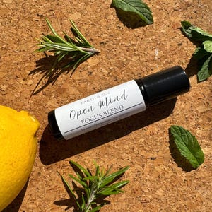 May include: A black essential oil roller bottle with a white label that reads "Open Mind FOCUS BLEND." The bottle is surrounded by fresh rosemary, mint leaves, and a yellow lemon on a corkboard surface. The product is from Earth & Zen.