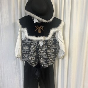 Peut inclure: Un costume de pirate pour enfant comprenant un chapeau noir bordé de blanc, une cape noire avec un emblème de tête de mort et de tibias croisés, un gilet à motifs, une chemise blanche et un pantalon noir.
