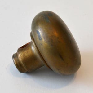 May include: A vintage brass door knob with an oval handle and a cylindrical base. The knob has a tarnished, aged appearance, suggesting it is an antique or a well-used item. The brass has a warm, golden hue.