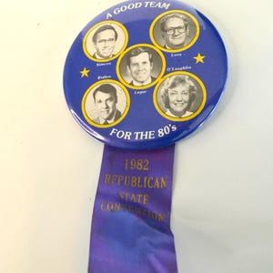 May include: A blue political campaign button with the text "A Good Team" and "For the 80's". It features portraits of five individuals and a purple ribbon with "1982 Republican State Convention" printed in gold.