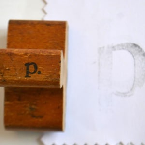 Könnte beinhalten: Ein Holzstempel mit dem Buchstaben "p" in schwarzer Tinte gedruckt. Der Stempel ist rechteckig und hat einen Griff. Ein schwacher Abdruck des Buchstabens "P" ist auf dem Papier neben dem Stempel zu sehen.