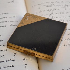 May include: A black and gold compact with a hinged lid. The compact has a black enamel surface with a gold trim. The compact is closed.