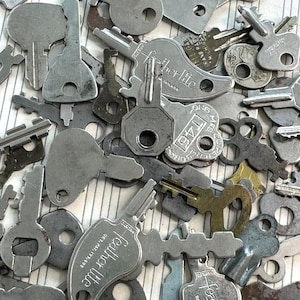 May include: A collection of various vintage metal keys in different shapes and sizes. Some keys have the brand name "Featherlite" stamped on them. The keys are scattered on a white and light gray striped surface.