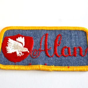 May include: Embroidered patch with the name "Alan" in red script. The patch features a blue background, a red shield with a white bird emblem, and a yellow border.