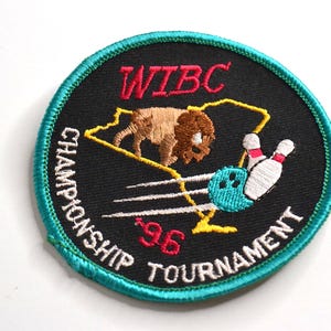 Könnte beinhalten: Ein runder gestickter Aufnäher mit schwarzem Hintergrund und türkisfarbenem Rand. Er zeigt einen braunen Büffel, eine blaue Bowlingkugel und weiße Bowlingkegel. Der Text lautet WIBC CHAMPIONSHIP TOURNAMENT 96 in rotem und weißem Garn.