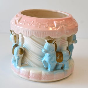 Könnte beinhalten: Ein Keramik-Karussell-Pflanzgefäß in Pastellrosa, Blau und Weiß. Der Pflanztopf hat ein Karussell-Design mit blauen Pferden, goldenen Akzenten und einem rosa Baldachin. Der Sockel und der obere Rand sind ebenfalls rosa.