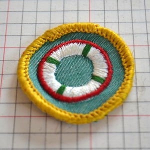 May include: Embroidered patch featuring a life preserver design. The patch has a teal background with a white and red life preserver in the center, accented with green stripes. A yellow embroidered border frames the design.