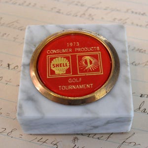 May include: A square, marble-like paperweight with a red and gold circular emblem. The emblem reads "1973 Consumer Products Shell Golf Tournament". The Shell logo and a stylized tree are visible. The paperweight is a vintage collectible.