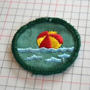 May include: Embroidered patch featuring a beach ball in red and yellow, floating on blue waves. The patch has a green background and a green stitched border. A fun, colorful design for clothing or accessories.