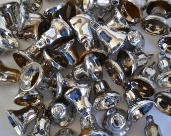 May include: A pile of silver-colored bells, some with a slight gold tint, are scattered on a white surface. The bells are all different sizes and shapes, but they all have a similar design.