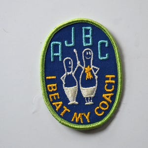 Puede incluir: Un parche bordado azul y verde con el texto "AJBC I BEAT MY COACH" y una imagen de dibujos animados de dos bolos.