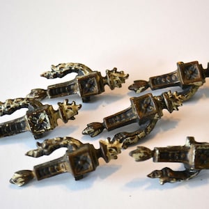 May include: Five antique bronze drawer pulls with ornate detailing. Each pull features a central square element with a floral design, flanked by curved arms and decorative end pieces. The pulls have a distressed, aged appearance.