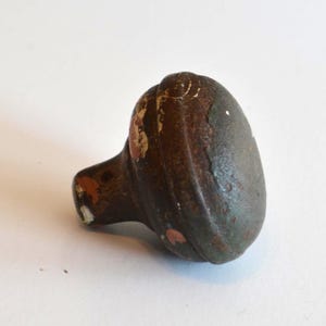 May include: A round, mushroom-shaped metal cabinet knob with a dark, weathered brown and grey finish. The surface shows signs of wear with patches of chipped paint revealing a reddish-brown base layer underneath the textured, aged exterior.