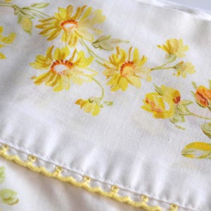 Op de afbeelding: Een witte stoffen textiel met een geprint patroon van gele en oranje bloemen met groene bladeren. De rand van de stof is afgewerkt met een decoratieve gele schulprand die kleine cirkelvormige uitsparingen langs de boord bevat.
