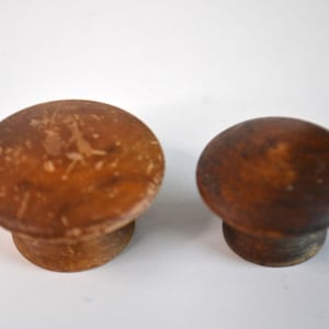 May include: Two round wooden knobs with a smooth, polished finish. The larger knob on the left features a light brown tone with visible surface scratches, while the smaller knob on the right is a darker, solid brown color with a uniform appearance.