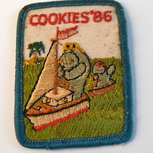 May include: Embroidered patch with the text "COOKIES '86". The patch features a sailboat with a flag and two cartoon characters in a grassy field. The patch has a blue border and is rectangular in shape.