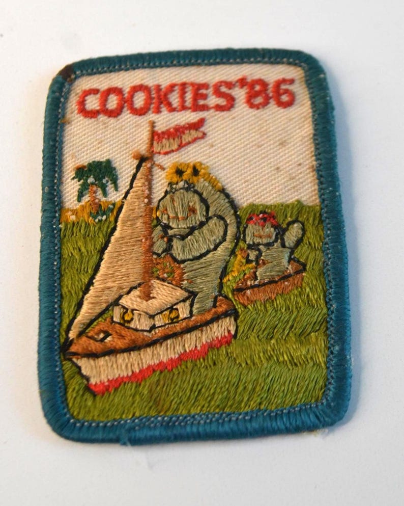 May include: Embroidered patch with the text "COOKIES '86". The patch features a sailboat with a flag and two cartoon characters in a grassy field. The patch has a blue border and is rectangular in shape.