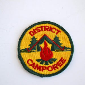 May include: A round, yellow embroidered patch with a green border. The patch features a red flame, a green tent, and two green trees. The text "DISTRICT CAMPOREE" is embroidered in red.